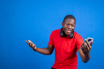 Joyful young man wearing headphones and using his smartphone against a vibrant blue background....
