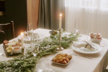 High angle shot of lit candles in silver candlestick on table covered with snow white tablecloth with pine garland, plates with food on top