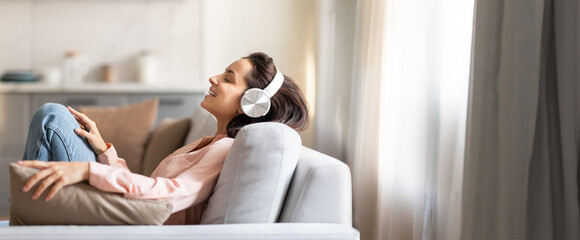 A woman enjoys her leisure time on a comfortable sofa, wearing headphones and relaxing. Soft natural light fills the cozy living room, creating a serene atmosphere.