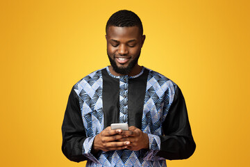 Smiling black guy using modern smartphone, wearing african t-shirt, pink studio background. African...