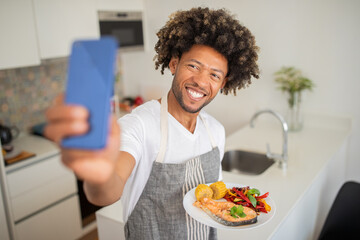A smiling black man is holding a plate of healthy food while taking a selfie in his cozy kitchen, showcasing his cooking skills and enjoying the moment. Vibrant meal features salmon and vegetables.