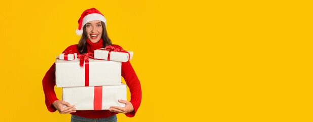 A cheerful woman in a red sweater and Santa hat joyfully carries wrapped Christmas presents. The bright yellow background adds to the festive atmosphere and holiday spirit, copy space