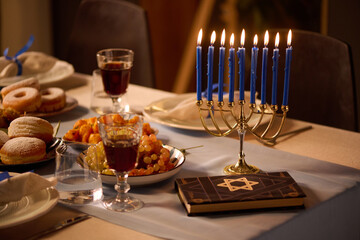 Medium close up of blue lit candles in golden candlestick, Torah with star of David on cover and different dishes on table top