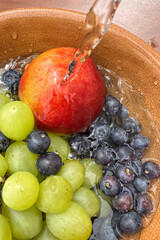 closeup of fresh fruits, blueberries, peach, and grapes,  in a bowl under running water while washing the fruit.
