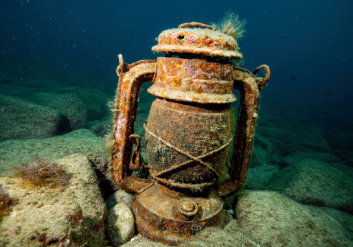 An ancient metal lantern rests on the ocean floor, covered in marine growth near rocky seabed in a picturesque underwater environment