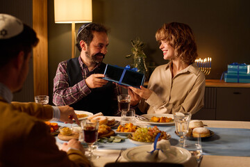 Jewish husband and wife exchanging gifts while celebrating Hanukkah with friend who enjoying traditional dish