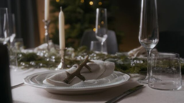 Close up view of festive dining table set with white plates, napkin adorned with antler holder, glassware, candles and evergreen centerpiece, with softly lit Christmas tree in background