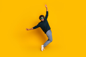 Young man in sunglasses joyfully posing on vibrant yellow background with arms raised upward...