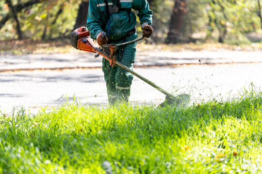 A landscaper using a string trimmer to maintain grass along a park pathway during a sunny autumn day in a public recreational area