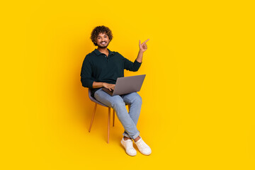 Young man with laptop on yellow background points enthusiastically. Student is seated wearing casual black shirt jeans