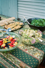 Film photo of a colorful outdoor table spread featuring a variety of fresh salads, a bowl of mixed fruit, bread, and savory pastries, set on a patterned tablecloth for a summer gathering.