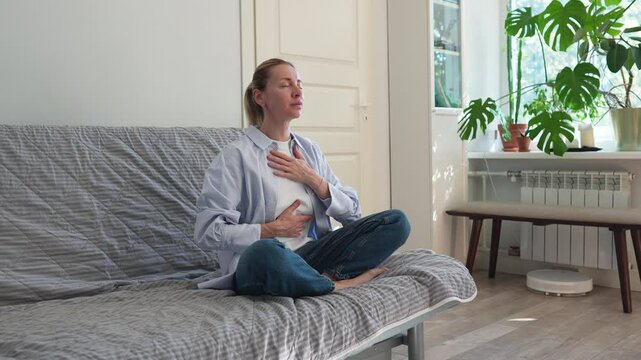 Calm woman meditating with controlled breathing on sofa, focus on deep breaths at home. Relaxation, self care, inner harmony, mental recharge, stress relief, meditation, self focus, time with yourself