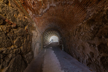 An arched brick tunnel with textured, aged walls leading towards an exit illuminated by bright daylight