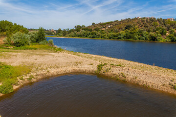 A serene riverside landscape with gently sloping sandy banks, lush greenery, and a calm river...