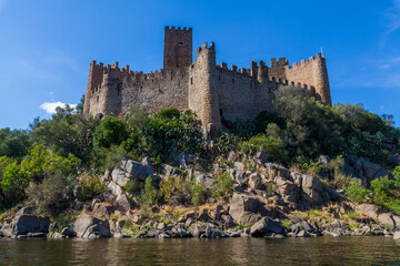 A historic castle stands majestically atop a rocky island surrounded by calm river waters, with a backdrop of lush hills