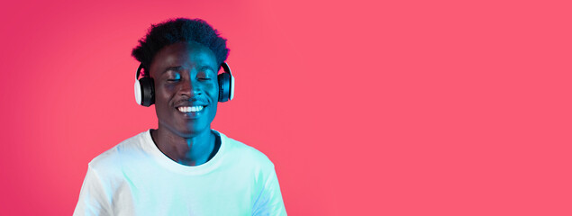 A young black man smiles while listening to music through headphones, showcasing his enjoyment. The bright pink backdrop adds a lively touch to the atmosphere, enhancing the moment.