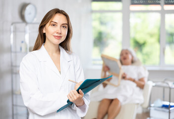 Obraz premium Smiling young female medical person standing at exam room of aesthetic center holding folder and pen in her hands