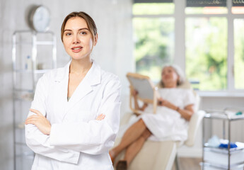 Fototapeta premium Smiling young lady wearing medical scrub standing at treatment room in front of female patient lying at examination couch.