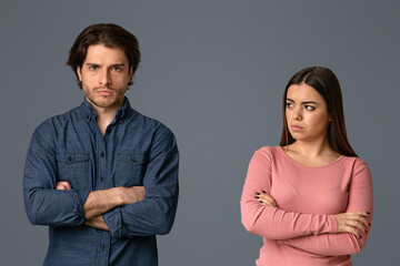 Young woman looking with sorrow at her offended husband after their argue, couple standing with folded arms over grey background, empty space