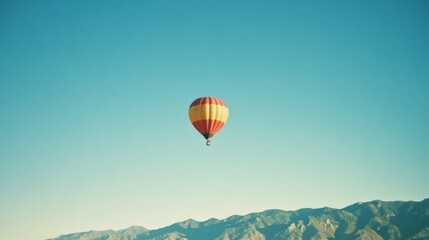 Fototapeta premium A lone hot air balloon drifts serenely against a clear blue sky, with a majestic mountain range providing a picturesque backdrop.