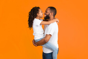 Happy Loving Family. Profile Side View Portrait Of Cheerful African American Man Holding Carrying Excited Daughter On His Hands, Having Fun Standing Isolated Over Yellow Studio Background