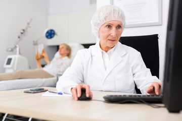 Positive elderly female cosmetologist working with laptop filling out medical form of patient before procedures in aesthetic medicine office