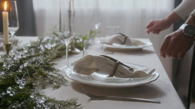 Close up view of hands of man placing napkins decorated with antler ring holders on plates while setting table for Christmas dinner at home