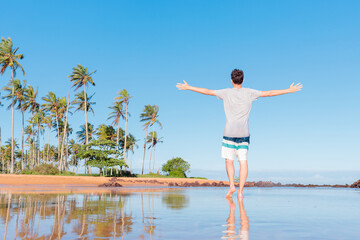 Man enjoying a tropical beach trip, standing barefoot with arms open by the water. Palm trees and a clear blue sky create the perfect scene for relaxation and travel