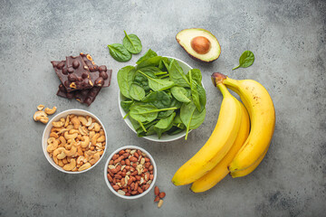 Assorted magnesium rich foods like bananas, spinach, peanuts, cashews, and dark chocolate on a gray...