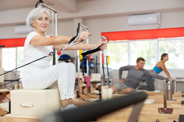 Senior woman works out on in gym on Pilates simulator. Sports club client on reformer with cable...
