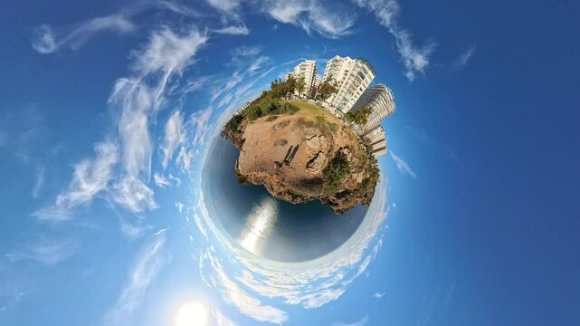 A 360-degree tiny planet effect image capturing the rotation of a rocky coastal landscape with buildings, greenery, and an expansive blue sky, creating a surreal, spherical world perspective