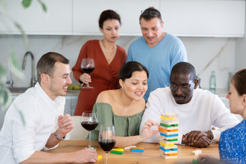 Joyful friends in their middle years sitting around table playing brick game and drinking wine in goblets