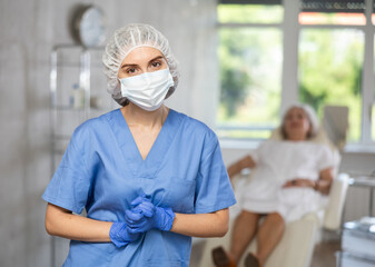 Obraz premium Portrait of young female doctor wearing blue uniform, medical mask and gloves standing in treatment room at beauty clinic