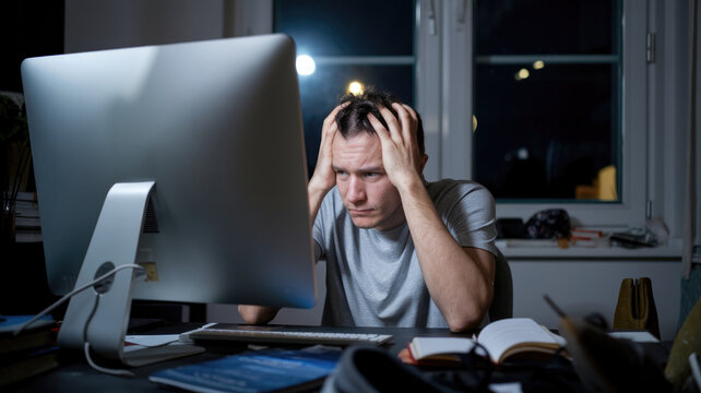 A man is sitting at a desk with a computer monitor in front of him. He is looking at the screen with a look of frustration on his face