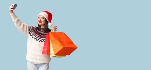 Pretty young woman in Santa hat taking selfie with shopping bags on light blue background with space for text