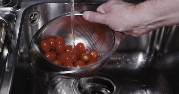 Ein Mann w&auml;scht Tomaten in einem Sieb aus Edelstahl unter flie&szlig;endem Wasser.