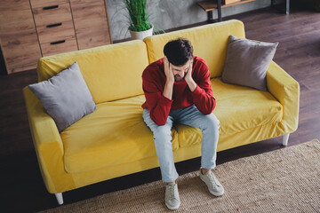 Obraz premium Young man in casual pullover sitting on yellow sofa in comfortable apartment enjoying a relaxed weekend day indoors with natural daylight