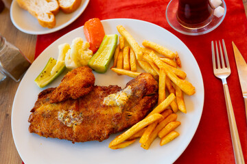 Portion of Turkish fried fish fillet with potato and vegetables served on plate in restaurant with mug of tea.