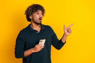 Young man in black shirt holding phone, pointing with amazement against vibrant yellow background,...
