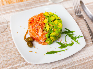 Fresh salmon tartare with diced avocado sprinkled with sesame seeds served with arugula leaves and pickled capers on white plate