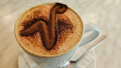 White coffee cup and saucer with cappuccino latte chocolate powder in the shape of the letter 'v' against a marble table, including silver spoon and napkin