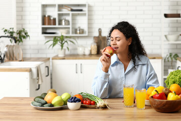 Young happy African-American woman with glasses of juice, different vegetables and fruits in kitchen. Independence from Meat Day