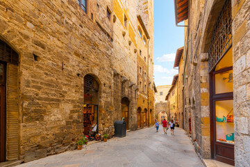 Fototapeta premium Tourists walk through a narrow stone street of cafes and shops in the historic center of the Tuscan Hill Town of San Gimignano, Italy.