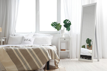 Interior of light bedroom with cozy bed and monstera leaves on bedside table