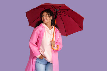 Happy young African-American woman in raincoat with umbrella on lilac background