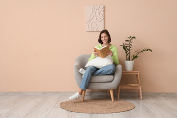 Pretty young woman reading book on armchair near beige wall