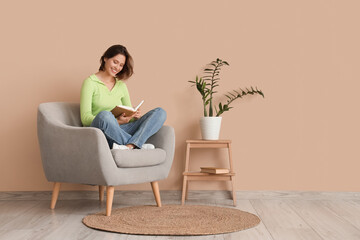 Pretty young woman reading book on armchair near beige wall