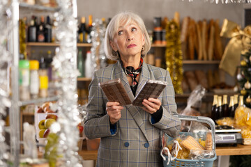Elderly woman buyer chooses sweet chocolate in christmas decorated grocery store