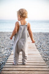 Beautiful toddler in summer grey jumpsuit stands alone on path leading to the seashore on warm day.