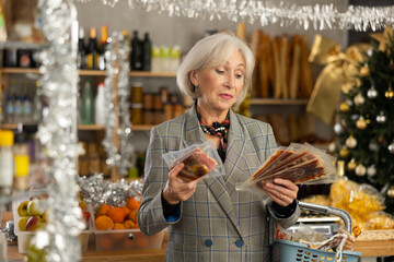 Elderly woman buyer chooses packaged jamon in christmas decorated grocery store